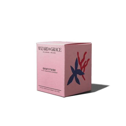 Studio shot of the pink cardboard box for the Wizard &amp; Grace Gratitude candle set against a white background. The front displays "Wizard &amp; Grace Wellbeing Ireland," "Gratitude - Thyme, Frankincense &amp; Bergamot," and "Natural Essential Oil Candle - 20cl/160g," with an abstract dark blue flower and red coral-like branch graphic on the adjacent side.