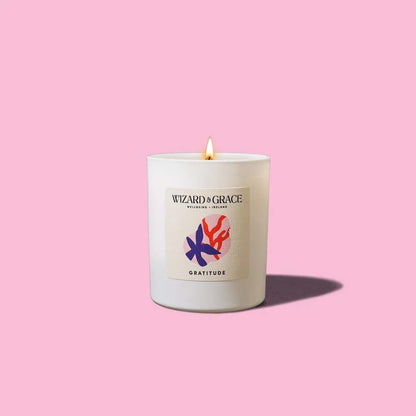 A white matte glass candle with a lit flame, displayed against a soft pink backdrop. The label displays the brand name "Wizard &amp; Grace Wellbeing Ireland," the scent name "Gratitude," and an artistic illustration of dark blue and red organic shapes.