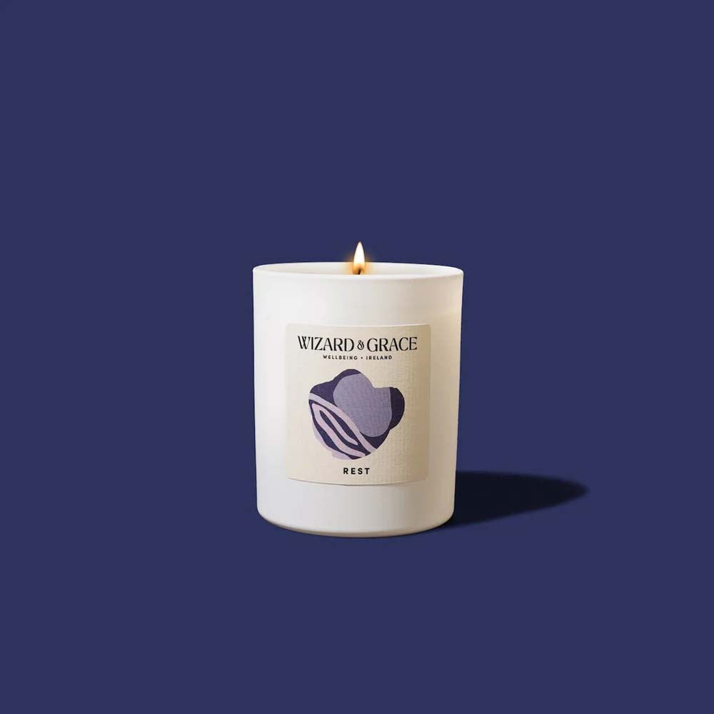 Lit Wizard &amp; Grace "REST" natural candle in a white jar set against a solid dark navy blue background.