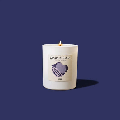 Lit Wizard &amp; Grace "REST" natural candle in a white jar set against a solid dark navy blue background.